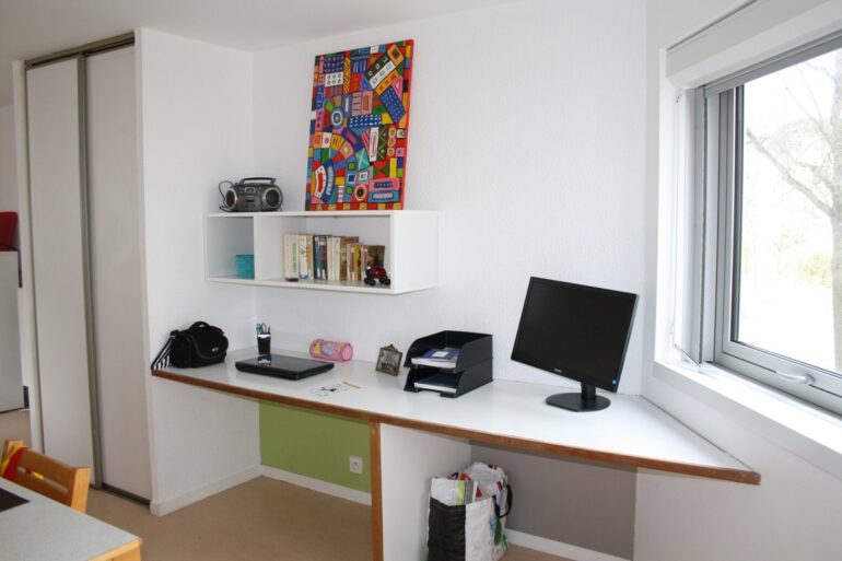 Chambre étudiante avec bureau blanc en L, moniteur Philips, étagère de livres et penderie, lumière naturelle